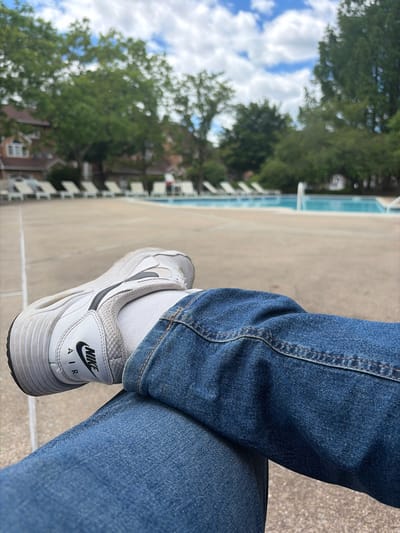 The image shows a person wearing blue jeans and white Nike Air sneakers sitting on a concrete surface. The person has their legs crossed. In the background, there are trees, buildings, and a blue sky with clouds. The location is likely outdoors near a building and greenery, but there is no concrete evidence to support it is within the city of Edison, United States of America. The photo appears to have been taken during daylight hours.