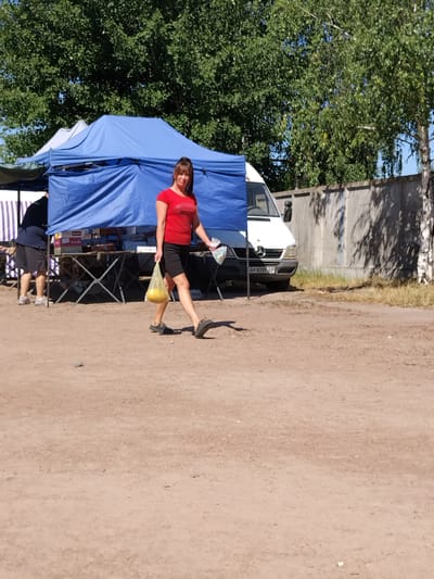 A woman with brown hair is walking past a blue tent at an outdoor market in Irshansk, Ukraine. She is wearing a red shirt and black shorts, and carrying a yellow plastic bag. In the background, a white van is parked next to the tent, and a grey concrete wall is visible. The ground is dirt and gravel.