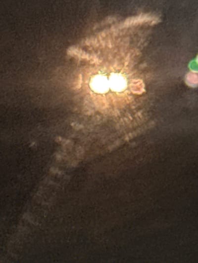A dark, textured surface dominates the scene. A bright, blurred light source appears as two overlapping circles, surrounded by a radial burst of light. A smaller, reddish-pink blurred shape sits slightly above the light. A faint, hazy trail of light stretches from the main light source towards the right side of the frame, suggesting movement or diffusion of light. A few other blurred, colored circles are present in the upper left corner.