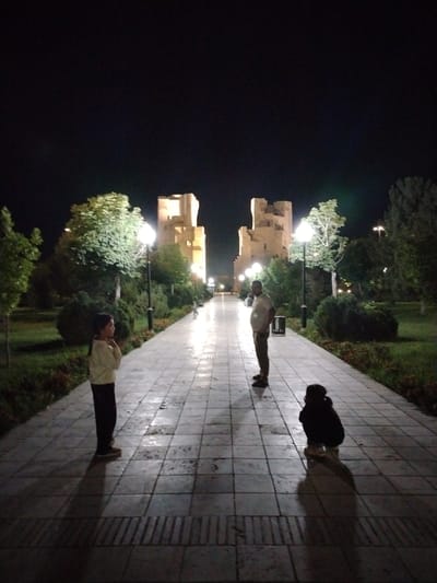The image depicts a night scene in Shahrisabz City, Uzbekistan. A stone pathway, illuminated by lampposts, stretches into the distance. Two large, ancient-looking stone structures, possibly fortresses or palaces, are visible in the background, lit from below. In the foreground, three individuals are present. A young girl stands on the left, facing away from the camera, wearing a light-colored top and dark pants. Further down the path, a man stands facing left, dressed in a light t-shirt and dark pants. On the right side of the path, a person is crouched down, facing away from the camera, wearing dark clothing. Greenery and trees line the pathway on both sides, and the sky above is dark and clear, indicating nighttime.