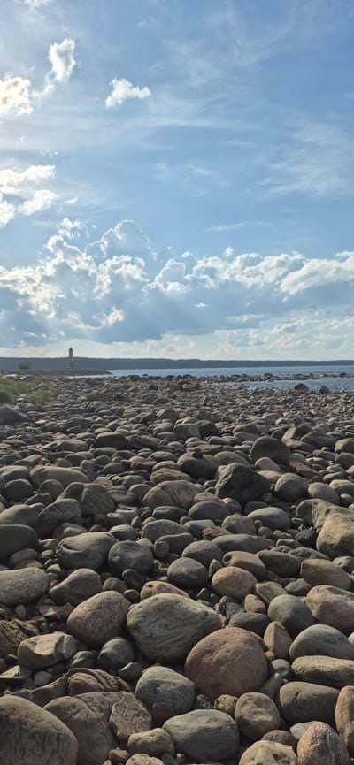 The image depicts a rocky shore with a body of water in the background, under a cloudy sky. A lighthouse and a small building are visible on the horizon, situated on a rocky peninsula. The foreground is dominated by a dense arrangement of smooth, rounded stones of various sizes and colors. The water appears calm, and a line of trees forms the edge of the land beyond the water. The overall scene suggests a coastal or lakeside environment.