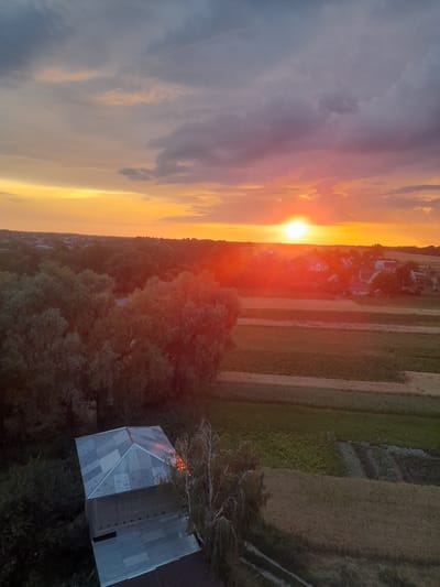 An aerial drone shot captures a sunset over the outskirts of Borodianka, Ukraine. The sky is a dramatic canvas of oranges, yellows, and purples, with thick clouds streaking across the horizon. The sun, a brilliant orb, is low in the sky, casting a warm glow over the landscape. In the foreground, a dilapidated, corrugated metal-roofed structure is visible, with some evidence of damage or disrepair. The surrounding area is a mix of trees and agricultural fields, with a small village nestled in the middle distance. The overall impression is one of a rural setting bathed in the warm, fading light of dusk, with a sense of quiet and possibly abandonment.
