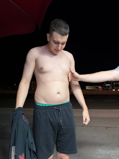 A young man with short dark hair is standing outdoors at night, looking down. He is shirtless, wearing black shorts with a green waistband. A black shirt is draped over his left arm. A hand is seen pressing into his right side. In the background, a road is visible with lights from vehicles and buildings. The scene appears to be near an open area, possibly a parking lot or roadside rest stop.