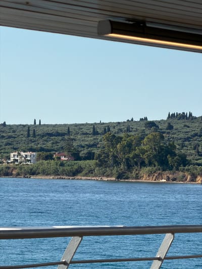 The image depicts a coastal landscape in the Municipal Unit of Kastro - Kyllini, Greece. A clear blue sky dominates the upper portion of the frame. In the middle ground, a lush green hillside covered with dense vegetation and scattered cypress trees slopes down towards the water. Two houses are visible on the hillside; one is a modern, multi-story white building with multiple windows, and the other is a smaller, reddish-tiled roof structure. The foreground features a body of calm blue water. A metal railing, part of a viewing platform or walkway, runs horizontally across the bottom of the image, partially obscuring the view of the water. The overall scene is serene and highlights the natural beauty of the coastal region.