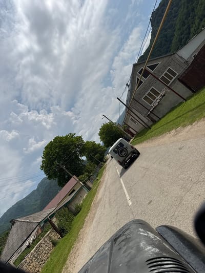 The image captures a street scene in Каменномостское сельское поселение, Russia, viewed from a vehicle. A white SUV is driving on a paved road. The road has a white dividing line. On the right side of the road, there is a two-story building constructed of light-colored brick, with several windows and triangular attic windows. Adjacent to this building is a grassy area and a brown fence. On the left side of the road, there is another building with a red metal roof and a stone wall at its base. Behind the buildings, a lush green mountain covered in trees rises steeply. The sky is filled with dramatic white and gray clouds. The image is taken at an angle, as if the photographer is looking out from a moving vehicle.