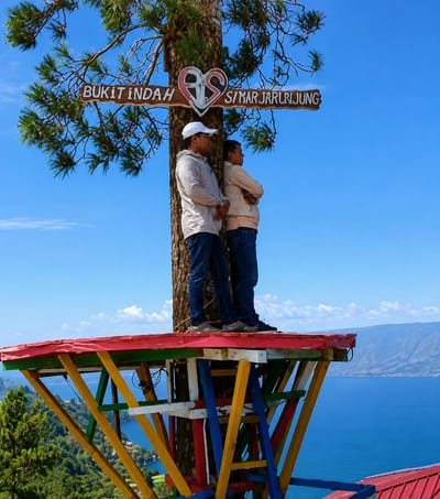 In Parik Sabungan, Indonesia, two individuals stand on a vibrant, multi-colored wooden platform built around a tree trunk. The man on the left wears a white cap, a light gray hoodie, and dark blue jeans. The man on the right, partially obscured by the first, is wearing a light beige hoodie and dark blue jeans. Both men are looking towards the right, with arms crossed or clasped in front of them. A wooden sign is attached to the tree trunk above them, bearing the text "BUKIT INDAH" on the left and "SIKAR JARRLLJUNG" on the right, with a heart symbol in between. The platform overlooks a vast, blue body of water, likely a lake, with lush green hills and mountains in the background under a clear blue sky with scattered white clouds. The bright, natural lighting suggests it is daytime.