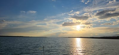 The image depicts a wide expanse of calm water, likely a lake or reservoir, with a low, tree-lined horizon in the distance. The sky is a dominant feature, filled with scattered clouds that are illuminated by the setting sun. The sun itself is partially obscured by clouds, casting a golden glow and creating a shimmering reflection on the water's surface. The overall scene conveys a peaceful and serene atmosphere. It is plausible that this scene is located near Pristan'-Przheval'sk, Kyrgyzstan, given the natural landscape and potential for such bodies of water in the region. There are no people or specific actions visible in the image; the focus is on the natural environment.