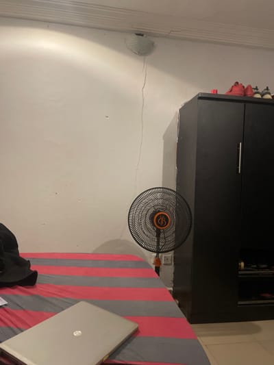 A room interior in Ogba, Nigeria, features a black cabinet mounted on a white wall. A black electric fan with an orange center is positioned near the cabinet. Adjacent to the fan, a bed with red and gray striped bedding holds a silver laptop. A black garment is partially visible at the bottom of the bed. A white vent is visible on the left wall, and a crack runs horizontally across the wall surface.
