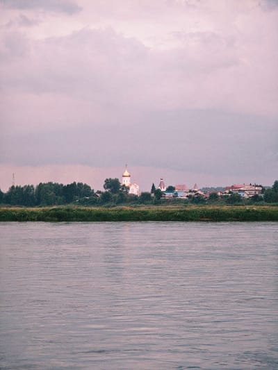 The image shows a body of water in the foreground with a town or village on the far bank. The sky is overcast and a pale pinkish-purple color. On the far bank, there is a line of trees, behind which are buildings with red roofs, and at least one white building with a golden dome, likely a church. The water is calm with visible ripples. The scene appears to be a landscape view, possibly from a boat or the opposite riverbank. The town is situated along a tree line, with some structures visible beyond the trees.