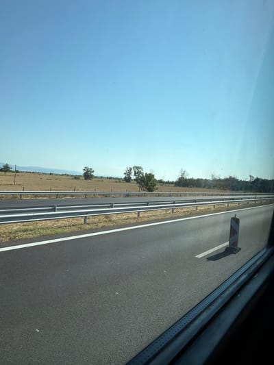 This is a horizontal, eye-level, medium shot of a highway in Kolarovo, Bulgaria. The road is paved and has a white line painted on the right side. There are two guardrails on the left side of the road, one above the other. Beyond the guardrails, there is a dry, grassy field with some scattered trees. In the distance, there are hills. The sky is clear and blue. There is a striped traffic cone on the right side of the road. The photo appears to be taken from a vehicle, as indicated by the reflection on the right side of the frame.