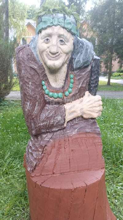 This media file is a vertical, close-up photograph of a wooden sculpture of a woman's head and upper torso carved from a log. The sculpture is painted. The woman has a wrinkled face, smiling with her mouth open, and appears to be wearing a green headdress adorned with leaves. She has dark eyes and her hair is grey. She is wearing a dark brown garment with a textured pattern, and a necklace of round, green beads. One of her hands, carved from wood and painted a lighter shade, is visible grasping the garment or log. The sculpture is set against a backdrop of green grass with some small white flowers and blurred trees and a building in the distance. The lighting suggests it is daytime, possibly overcast, given the soft light and slight haze. The overall impression is of a whimsical or folkloric figure. No visible text is present in the image. The location context provided is Khmilnyk, Ukraine.