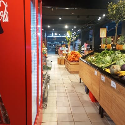 This image depicts the interior of a supermarket or grocery store in Kisa Central ward, Kenya, focusing on a produce aisle. The scene is well-lit with numerous recessed ceiling lights in a dark overhead panel. On the far left, a large red Coca-Cola branded refrigeration unit is partially visible. Adjacent to it, a silver shopping cart with a green basket is positioned. The floor is covered with light-colored square tiles.
To the right, a prominent display of fresh produce is arranged in multiple elevated wooden bins. These bins contain various items including large green cabbages, assorted leafy greens, pumpkins, and bananas. Further down the aisle, a bin is filled with a large quantity of red tomatoes. Above the produce section, pineapples are displayed on an upper shelf, and decorative artificial trees or potted plants, including one resembling a banana plant, are interspersed.
In the mid-ground, shelves stocked with various packaged goods and bottles are visible extending into the background. A partially obscured black sign with white text, possibly reading "DIMES BICYCLICS," is visible above one of the distant aisles. No individuals are present or engaged in any actions within the frame. Security cameras are visible mounted on the ceiling.