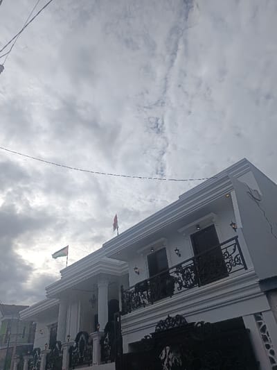 This image depicts a two-story, predominantly white building situated in Sindangasih, Indonesia, viewed from a low-angle perspective. The building exhibits a modern architectural style with classical influences, featuring decorative moldings along the roofline and window surrounds. A prominent second-floor balcony with an ornate black wrought-iron railing extends across a significant portion of the facade. Multiple wall-mounted light fixtures are visible on both levels of the building. Two flags are displayed on the roofline: the flag of Palestine (black, white, green horizontal stripes with a red triangle at the hoist) is positioned on the left, and the flag of Indonesia (red over white horizontal bi-color) is positioned on the right. The sky is overcast, displaying various shades of grey and white clouds. Several thin, dark utility lines traverse the upper left portion of the frame. A section of an ornate dark-colored gate is visible in the lower right foreground, partially obscuring the ground level. No individuals, actions, or interactions are visible within the scene.