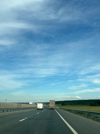 The image depicts a highway in Malaya Tsarevschina, Russia. Several vehicles, including trucks and a van, are traveling on the roadway. The sky is a light blue with wispy clouds. To the right of the highway is a green field and a treeline. The road has a concrete barrier on the left and a guardrail on the right. White lane markings are visible on the road surface.
