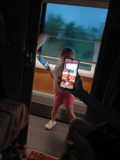 In a dimly lit train compartment, a young girl in pink shorts and a white t-shirt stands by the window, holding a tablet. Her back is to the viewer, and she appears to be looking out at the passing scenery. In the foreground, a hand holds a smartphone, its screen displaying a live camera feed of the girl. The phone's screen shows the girl in a red top and pink shorts, with a blurred background suggesting motion. The compartment's interior is visible, with luggage draped over a seat and a rug on the floor. The window shows a hazy, blueish sky and green trees, indicating that the train is in motion, possibly traveling through the countryside near NovoYa kupovo, Russia.