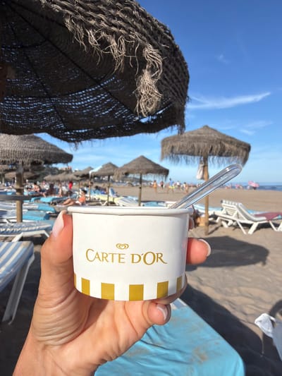 The image shows a close-up of a hand holding a white paper cup with a "Carte d'Or" logo and yellow stripes. A clear plastic spoon is inserted into the cup. In the background, a beach scene is visible with straw umbrellas, lounge chairs, and people relaxing. The ocean and a blue sky with some clouds are also present. The location is identified as the beach in Benicàssim, Spain. The overall atmosphere is one of leisure and enjoyment on a sunny day.