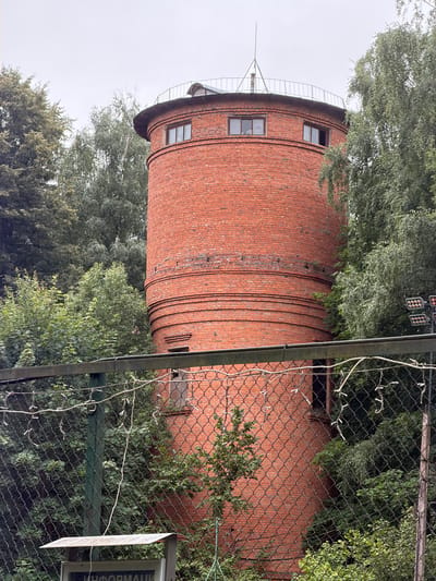 The image shows a red brick tower, likely a water tower, in Novo-daryino RAN, Russia. The tower has a cylindrical base with decorative horizontal bands and a wider top section with several small windows. A metal railing encircles the top of the tower, and a thin antenna or spire rises from the center. The tower is surrounded by lush green trees and foliage, suggesting a natural or park-like setting. In the foreground, a chain-link fence partially obscures the view of the tower's base. There are strings of small white lights draped over the fence and a sign with Russian text, possibly "ИНФОРМАЦИИ" (information), visible below the fence. The sky is overcast and grey, indicating a cloudy day. No people or animals are visible in the image.