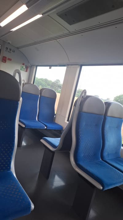 The image shows the interior of a train carriage, likely a public transport vehicle. There are several rows of blue seats with grey headrests arranged in facing pairs. The seats have a textured pattern. The floor is dark grey and reflects some light. Above the seats, the ceiling is white with visible lighting fixtures and what appears to be a ventilation grate. Large windows on the side offer a view of greenery and trees, suggesting an outdoor environment. There are some signs on the wall near the window, including one with "VT 653" and icons for Wi-Fi and a camera. The overall impression is a clean and modern train interior. The location of Quickborn, Germany, is not directly evident from the image itself but can be inferred from external information.