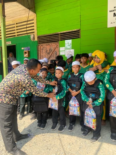 In Mekar Raya, Indonesia, a group of children wearing green and yellow uniforms and white skullcaps are gathered outside a building with bright green wooden walls. A man in a floral shirt is handing out plastic bags filled with school supplies to the children. Some children are wearing black vests with "YBM PLN" printed on them. The ground is covered with gravel, and there is a shadow of a person's leg and a purple object on the ground. A sign on the building reads "JL. A. Y." and a meter is visible on the wall.