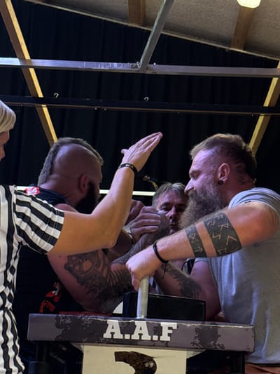 This image depicts an arm wrestling match in an indoor setting in Reisenberg, Austria. Two adult males are engaged in the competition at a specialized arm wrestling table, which bears the white letters "A.A.F" on its side. Both competitors have extensive tattoos on their arms, visible beards, and short or styled hair. The individual on the left side of the table wears a black shirt with visible tattoos, including a large design on his bicep, and has a mohawk-style haircut. The individual on the right wears a light-colored t-shirt, has a prominent beard, ear gauges, and a band of runic-style tattoos around his forearm. He grips a vertical handle on the table with his left hand.
A referee, partially visible on the far left, wears a black and white striped shirt and has light-colored hair; their right hand is raised above the competitors' locked hands, indicating an active phase of the match or a command. Another male, with graying hair, is visible behind and between the two competitors, observing the match intently. The background shows dark fabric or curtains, structural metal beams, and overhead lighting, typical of an event hall or gym.