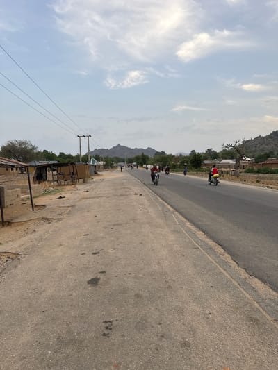 The image, oriented vertically with the sky on the left and the road on the right, depicts a rural or semi-urban road scene in Dawaki, Nigeria. The main subject is an asphalt road stretching into the distance, flanked by dusty shoulders.
In the midground, several individuals are visible riding motorbikes on the road. One person wearing a red shirt and a yellow vest is prominent, riding a motorbike towards the upper right of the frame (away from the viewer). Further down the road, other motorbikes and people can be seen, though less distinctly.
Along the right side of the road, various structures are present. Closer to the foreground, a utility pole with power lines runs parallel to the road. Below the power lines, there are rudimentary wooden structures, possibly makeshift stalls or small buildings, and some concrete blocks on the ground. Sparse vegetation and trees dot the landscape beside the road.
In the background, distant, hazy mountains rise against the horizon. The sky, occupying a significant portion of the image, is light blue with scattered white, wispy clouds, suggesting a partly cloudy day. The overall impression is of a warm, daytime setting. There is no visible text in the image.