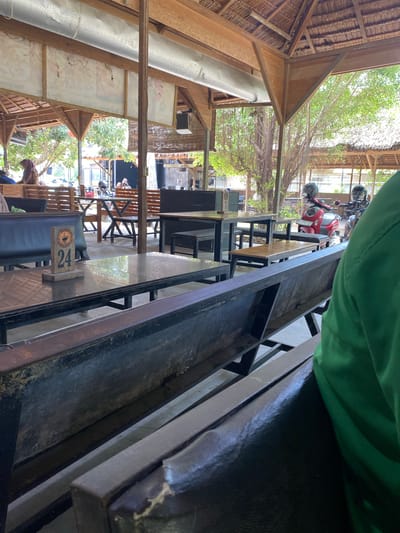 The image depicts an outdoor seating area, potentially a cafe or restaurant, in Gampongaceh, Indonesia. The structure features a thatched roof supported by wooden beams and pillars. Tables and benches are visible, constructed of metal and wood. Table number "24" is displayed. Several motorcycles are parked in the distance. The environment suggests a tropical or semi-tropical climate, with visible greenery.