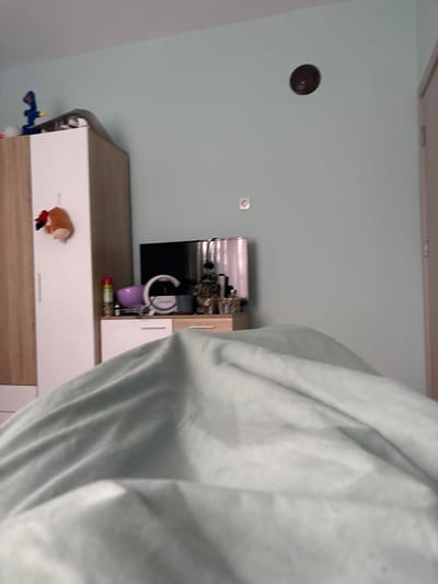 The image shows the interior of a room. In the left background, there is a light blue wall with a circular vent and an electrical outlet. A television is mounted on a cabinet with drawers and shelves. A white blanket dominates the right side of the image, obscuring the view. There are no people visible. The location, Nadarevo, Bulgaria, cannot be verified from the image.