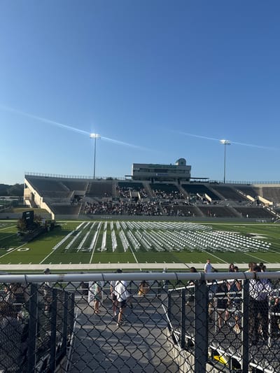 The image shows a stadium on a sunny day in Southlake, Texas, United States. The stadium is filled with rows of white folding chairs on the field and spectators in the bleachers. The stadium's press box is visible, with "DRAGON STADIUM" written on it. A metal chain-link fence obscures a portion of the view, with a few people standing behind it. Tall stadium lights stand on either side of the stadium. The sky is a clear, light blue.