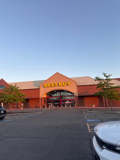 The image depicts the exterior of a Best Buy store in Bloomingdale, USA. The building has a brick facade with a distinctive triangular entrance. The yellow "BEST BUY" sign is prominently displayed. A parking lot is visible in front of the store, with a white car partially visible on the right edge. Trees are planted near the building. The sky is blue. There are people visible inside the store near the entrance. A handicap parking sign is visible in one of the parking spots.