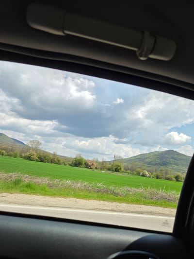 The view from inside a vehicle shows a landscape in Dobrodan, Bulgaria. A green field stretches towards a mountain covered in vegetation. A road runs along the edge of the field. The sky is cloudy with patches of blue. The interior of the vehicle includes a white grab handle.