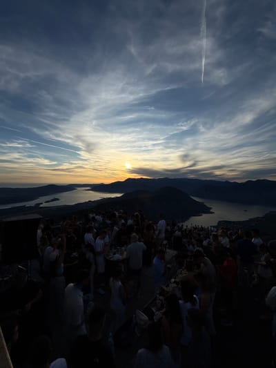 This is a wide-angle, high-angle photograph taken at sunset above the city of Špiljari, Montenegro. The scene depicts a lively outdoor gathering of a large group of people situated on a clifftop overlooking a bay. The sky is a dramatic mix of deep blues and oranges, with wispy clouds and a visible contrail from an aircraft. The sun is low on the horizon, casting a warm glow over the landscape.
In the foreground, a crowd of people are gathered. Many are dressed in casual summer clothing. Some are standing and talking, while others appear to be dancing or socializing around tables. There are drinks and possibly food visible on some surfaces. The atmosphere appears festive.
Beyond the gathering, a body of water, likely the Bay of Kotor, stretches out, surrounded by dark, silhouetted mountains that define the coastline of Špiljari and its environs. The lights of the city are faintly visible in the distance along the shoreline. The overall impression is of a celebratory event taking place in a stunning natural setting as day turns to night.