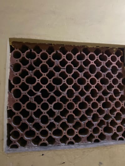 A close-up shot depicts a square window or vent covered with a lattice-like structure in Sabo, Nigeria. The lattice is constructed from a reddish-brown material, possibly terracotta or brick, arranged in a repeating pattern of interconnected circles and octagons. The background visible through the lattice is dark, suggesting limited light. The wall surrounding the window is painted a muted yellow or beige. The wall appears slightly worn.