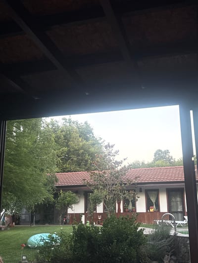 The image shows a backyard scene at a house in Prohod, Bulgaria. A single-story house with a red tile roof and white walls is visible. The house has several windows and a wooden exterior trim. In the foreground, there is a green lawn with some bushes and a small inflatable pool. A small red mushroom-shaped garden decoration is on the lawn. To the right, there is a lounge chair and a pool edge. The backyard is surrounded by lush green trees. The sky is overcast. The view is framed by a wooden overhang, suggesting the photo was taken from a porch or covered patio. There are no people visible in the image.