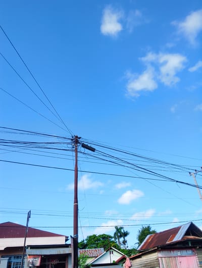 The image depicts a view of a street in Pompanua, Indonesia, characterized by a clear blue sky with scattered white clouds. Overhead, a dense network of power lines crisscrosses the frame, converging on a weathered wooden utility pole. The pole, situated in the mid-ground, is adorned with various insulators and electrical equipment, including a black rectangular box.

In the lower portion of the image, buildings are visible. On the left, a structure with a reddish-brown roof and white walls is partially visible, with windows that appear to be louvered. Below this, a smaller building with a corrugated metal roof is seen, displaying a sign with the text "FILTER BLACK". To the right of the utility pole, another building with a rusted, corrugated metal roof is prominent. This building features wooden walls and a pink facade, with a sign that reads "ADILLA CELLULAR". Trees, including what appears to be a palm tree, are interspersed among the buildings, adding a touch of greenery to the scene. The overall impression is one of a typical street scene in a tropical urban or semi-urban environment.