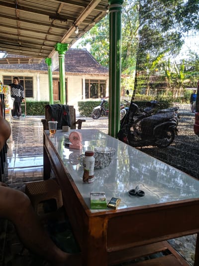 The image depicts an outdoor seating area, possibly a warung, in Ketoyan, Indonesia. A wooden table, covered with a reflective white surface, is laden with various items. These include a clear plastic cup with amber liquid, a small pink fan, a bottle of chocolate milk, a pack of cigarettes, a lighter, and a pair of scissors. The table is set in front of a low wooden structure with drawers. Behind the table, a person wearing a black and white zig-zag patterned shirt and dark pants stands near a display of products.
In the background, a building with a tiled roof and white walls is visible. Two motorcycles are parked on a gravel surface outside the seating area. A black mesh screen is draped over a green pillar, creating a semi-private enclosure. Lush green foliage and trees surround the scene, suggesting a tropical or sub-tropical environment. The lighting indicates it is daytime, with sunlight filtering through the trees.