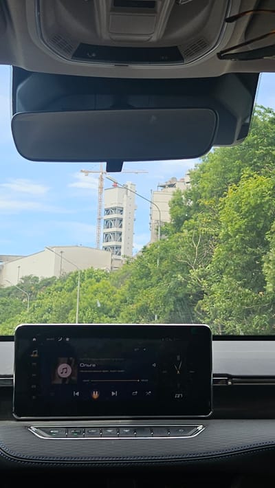 The image shows a view from inside a car, looking forward through the windshield. A car's infotainment screen is visible in the foreground, displaying a music player interface with the song title "Ольга". To the right of the screen, a clock face is displayed. Beyond the windshield, a bright, overcast sky is visible, with green trees and industrial buildings in the distance. A construction crane is prominent among the buildings. The scene suggests a temperate climate. No people are visible in the image. The location is identified as Verkhnebakanskiy, Russia.
