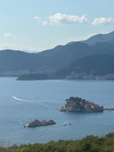 The image depicts a scenic coastal landscape, likely during the daytime under clear skies. On the left, a majestic mountain range slopes down towards the sea, with a town or city nestled along its foothills. The buildings appear densely packed, hinting at a populated area. White, fluffy clouds dot the bright blue sky above the mountains.
The right side of the image is dominated by the calm, blue sea. In the foreground, lush green trees line the coastline, suggesting a natural, verdant environment. Several islands are visible in the water, the most prominent being a small, rocky island adorned with quaint buildings topped with red roofs. A narrow causeway or bridge connects this island to the mainland, where more structures are visible. Another smaller, rocky islet lies nearby.
Several boats are present on the water. A few are moored near the island, while others are actively moving, leaving white wakes behind them. One boat, in particular, is creating a dramatic, arcing wake as it navigates the water. The overall impression is one of a picturesque and tranquil vacation destination. The location is identified as Blizikuće, Montenegro.
