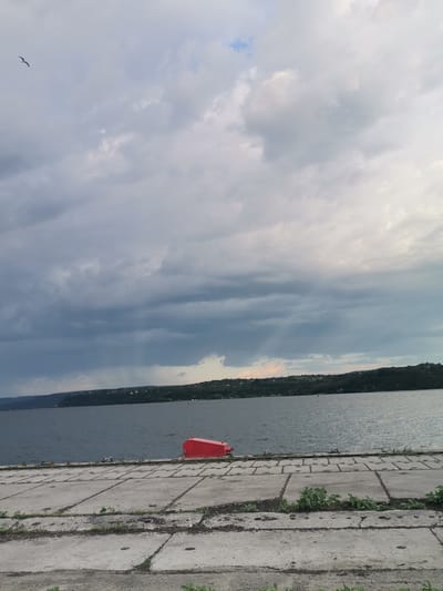 The image depicts a waterfront scene. In the foreground is a concrete pier or walkway, composed of rectangular slabs. Vegetation grows in the cracks. In the midground, there is a body of water, presumably a lake or river. A red object is floating in the water near the pier. In the background is a treeline. The sky is overcast with various shades of grey and some hints of blue. A bird is flying in the upper left of the image. Given the request for the location in Ezerovo, Bulgaria, it can be inferred that the waterbody is the lake, and the treeline corresponds to the surrounding natural landscape of the area.