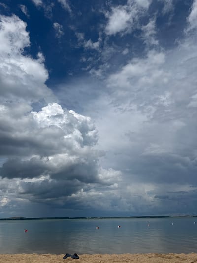 This media file is an outdoor, high-angle, wide shot of a beach and a body of water, likely a lake or a sea, under a dramatic sky filled with clouds. The image is oriented vertically, as if the photographer were holding the camera upright in portrait mode, but the scene is captured horizontally.

The foreground is a sandy beach, with a few pairs of dark shoes placed near the water's edge. The water itself appears calm and vast, stretching towards a distant, tree-lined horizon. Several buoys, marked with red and white squares, are dotted across the water, suggesting a designated swimming or boating area. The sky is a dynamic mix of deep blue in the upper left, transitioning to heavy, grey storm clouds that dominate the center and right portions of the frame. Sunlight breaks through some of the clouds, creating dramatic lighting.

The scene appears to be set during the daytime, likely late afternoon or early evening, given the dramatic cloud formations and the quality of light. The weather appears to be changing, with indications of an approaching storm. The location is identified as Володарский ауылдық округі, Kazakhstan.

The overall mood of the image is somewhat moody and awe-inspiring, due to the contrast between the calm water and the turbulent sky. There is no visible text in the image.