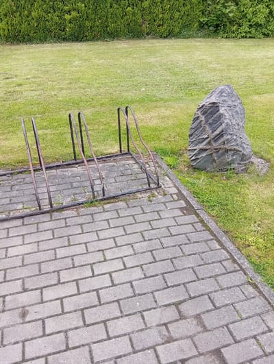 An outdoor scene depicts a paved walkway made of grey interlocking bricks. To the left of the walkway is a metal bicycle rack with four slots, showing signs of rust. Beyond the bicycle rack, a neatly trimmed green hedge forms a backdrop. To the right of the walkway, a large, dark grey stone sculpture with textured surfaces rests on a patch of grass. The area around the sculpture and extending further back is covered in green grass, with a few sparse patches of weeds and clover. The overall environment appears to be a public space, possibly a park or garden area, located in the city of Micaičiai, Lithuania.