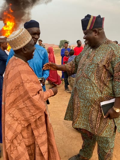 A group of people are gathered in an outdoor setting. Some individuals are formally dressed in traditional clothing. In the background, there is a large fire with thick black smoke. A man in patterned clothes extends his hand toward another individual. Several other people are seen observing the interaction and the surrounding environment. The setting appears to be a public space, potentially a gathering or event of some kind.