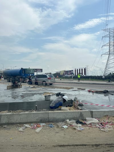 The image captures a daytime scene of a construction site alongside a road in Effurun, Nigeria, under a partly cloudy sky. In the foreground, a construction worker wearing a white hard hat and blue workwear is crouched, seemingly engaged in work on a concrete structure or curb. Beside them, there's debris, dirt, and visible red and white barrier tape.
Further into the scene, on the road, a large blue tanker truck with "JULIUS BERGER" prominently displayed on its side is visible, along with the number "B 7792". A silver SUV is also parked or moving nearby. Another worker, dressed in a yellow high-visibility vest, stands on the side of the road, close to several billboards.
In the mid-ground and background, multiple large electricity pylons or transmission towers are dominant features against the sky, carrying numerous power lines across the landscape. The billboards along the road display various advertisements; one shows text that appears to be "JUNIOR SAUTEE" and another has a "G-Money" logo. The overall setting depicts active infrastructure development or maintenance work in a semi-urban environment.