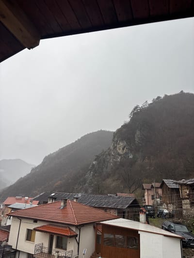 The image depicts an overcast day in Grohotno, Bulgaria, viewed from an elevated position looking down towards a residential area and distant mountains. A dark brown wooden overhang, possibly a roof eave or balcony ceiling, is present in the upper left foreground, with a visible water droplet at its edge. The sky is uniformly light grey, indicating heavy cloud cover or fog. The midground features a cluster of houses with various roof types, predominantly red clay tiles, but also dark corrugated metal and slate. One prominent light-colored, two-story house with a red-tiled roof and an ornate black wrought-iron balcony railing is central to the lower midground. A solar-powered light fixture is affixed to a pole near this house. Further back, a dark-colored BMW X5 SUV (partial license plate visible: CM606*) is parked on a grassy slope. Another dark SUV, identifiable by an Opel logo on its rear, is parked near a light pink residential building. Utility poles and power lines crisscross the scene between structures. Several older, weathered wooden buildings with dark roofs are visible on the right. In the background, steep, tree-covered mountains rise, partially obscured by mist or low clouds. The trees appear mostly bare, suggesting a dormant season. Rocky outcrops are visible on the closer mountain face. No individuals are visible.