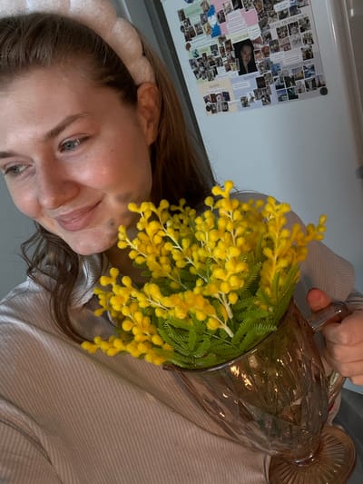 The image depicts a young adult female holding a clear, faceted glass pitcher containing a bouquet of yellow, fluffy flowers, consistent with mimosa. The woman, who appears to be taking a self-portrait, has fair skin, light-colored eyes, and light brown hair partially pulled back by a pale, textured headband. She wears a light-colored, vertically striped or ribbed collared shirt and exhibits a slight, downward-gazing smile. Her right hand is visible, gripping the handle of the pitcher.
The background reveals a light-colored refrigerator, prominently adorned with a collage of numerous small photographs and paper notes. Several notes display Cyrillic text, including "ЗДОРОВЬЕ" (Health), "СЕМЬЯ" (Family), "КАРЬЕРА" (Career), "ПУТЕШЕСТВИЯ" (Travels), "МЕЧТЫ" (Dreams), and "ДЕТИ, ТВОРЧЕСТВО" (Children, Creativity). A larger, central photograph of a woman's face is affixed to the refrigerator.
The scene takes place indoors, within a residential environment, likely a kitchen or a room with a refrigerator. The specific location within the city of Nikol'skii, Russia, cannot be determined from the visual evidence; however, the Cyrillic text confirms a Russian-speaking context. Lighting is soft and even, illuminating the subject and the immediate background.