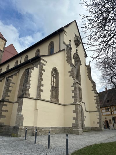 The image displays the exterior of St. Andreas Church in Weißenburg in Bayern, Germany, captured from a low angle under a partially cloudy sky. The main structure features cream-colored stucco walls, supported by prominent grey stone buttresses, and punctuated by multiple pointed-arch windows with intricate stone tracery. A dark gutter system runs along the eaves. A section of the main red-tiled roof is visible, with another red-tiled roof section appearing behind it to the left. A small, decorative stone corbel projects high from the right facade of the church. The foreground consists of a grey cobblestone plaza where four dark grey cylindrical bollards, each with a white reflective top band, are arranged. A small patch of green grass is visible at the lower right. Bare, dark tree branches extend into the upper right portion of the frame. In the distant background, to the right of the church, a traditional half-timbered house with white walls, dark timber frames, and a brown tiled roof is partially visible. Two indistinct human figures are present near the entrance of this background building, appearing to be standing or walking.