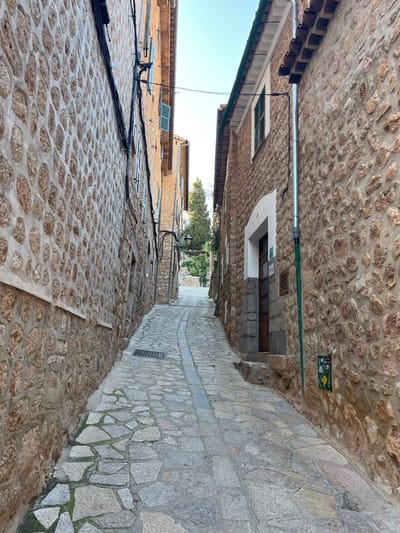 A narrow, stone-paved alleyway ascends through the city of Fornalutx, Spain, flanked by multi-story buildings with rough-hewn stone facades. The street curves gently to the right as it recedes uphill. The ground is paved with irregular flagstones, interrupted by a central strip of more uniform, rectangular pavers. A rectangular metal grate is visible on the left side of the paving.
On the left, the building wall is constructed from stacked, irregular, light brown stones, with black electrical cables running along its surface. Several windows with open light-blue louvered shutters are visible. The lower section of this wall has a smoother, lighter-colored plaster border around the stone.
On the right, another building features a similar stone facade. A prominent silver-colored metal downspout runs vertically along the wall. Below it, a small, dark green utility box with a white and yellow decal is affixed. Further up, a window with a closed dark green louvered shutter is present. A dark brown wooden door, set within an arched white frame, provides access to the building and is preceded by two shallow stone steps. The building's roofline shows visible terracotta tiles.
No people are present. A streetlight extends from the left building wall halfway up the alley. In the distant background, a tall, dark green coniferous tree is visible against a clear, light blue sky, above a higher stone structure.