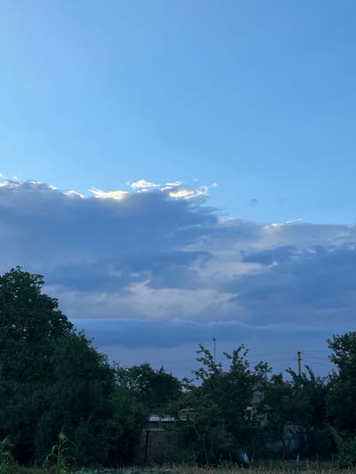 The image depicts a rural landscape under a partly cloudy sky. The sky is predominantly a bright blue, with large, diffused grey clouds present. Sunlight pierces through the clouds in some areas, creating illuminated patches. In the foreground, a dense line of dark green trees stretches across the lower portion of the image, obscuring much of what lies behind them. Some taller trees stand out amongst the foliage. A small section of a building with a tiled roof is visible behind the trees on the right side of the frame. In the far distance, a utility pole with wires is visible. The overall impression is of a tranquil, natural setting. The image was taken in the village of Samiilove, Ukraine.