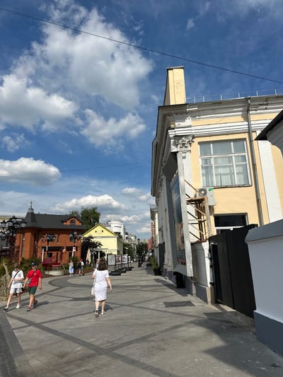 This is an eye-level, wide shot of a pedestrian walkway in Putyatino (Semivragovskaya S/A), Russia, under a bright, cloudy sky. The walkway is paved with grey interlocking stones, with darker grey rectangular patterns. On the left, several people are walking away from the camera. Two young males, one in a white t-shirt and shorts, and another in a red t-shirt and green shorts, are walking side-by-side. Further ahead, a woman in a white dress and a white purse is walking. Beyond them, a row of buildings lines the walkway. The building closest on the left is a reddish-orange structure with a dark pitched roof and multiple ornate black lampposts. To its right is a smaller, bright yellow building with a pitched roof. On the right side of the walkway is a large, pale yellow building with white architectural details and a prominent chimney. A large window with white curtains is visible on this building, and an air conditioning unit is mounted below it. A black fence is positioned in front of the right side of this building. In the distance, more buildings and trees are visible, along with streetlights and various signs. The overall atmosphere is that of a sunny day in a European-style town.