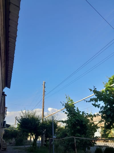 The image depicts an outdoor scene, likely a backyard or a street with residential buildings. The sky is clear and blue, with a few wispy clouds visible towards the horizon. In the foreground, on the left, a portion of a building's corrugated metal roof is visible, extending from the top left corner downwards. The building itself appears to be constructed with stone or concrete.

Dominating the middle ground is a tall, wooden utility pole with several wires extending from it. The pole is weathered and has insulators attached. To the right of the pole, a trellised vine with lush green leaves is prominent, with some branches reaching upwards. Several other trees and green foliage are scattered throughout the scene, suggesting a somewhat vegetated area. A wire fence runs horizontally across the lower part of the image, separating the foreground from the background.

In the background, residential buildings with visible windows and tiled roofs are present. The lighting suggests it is daytime, with the sun casting shadows. The overall impression is that of a somewhat rural or suburban setting within the city of Artashat, Armenia, characterized by a mix of natural elements and man-made infrastructure. No people are visible in the image.