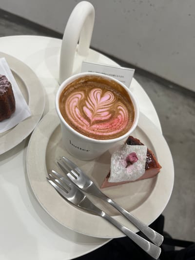 A top-down, medium-shot image shows a white table in Nakano, Japan, with a latte and dessert. A white paper cup with the word "butter" printed on it holds a latte featuring pink latte art in a fern pattern. A white plate holds a slice of cake, topped with white mochi and a pink cherry blossom. Two silver forks are placed diagonally on the plate. Another small white plate with a dark brown pastry is in the lower left corner. A piece of paper is partially visible next to the coffee cup. The table has a white, rounded edge. A gray wall or floor is visible in the background, along with a dark shape that may be clothing.